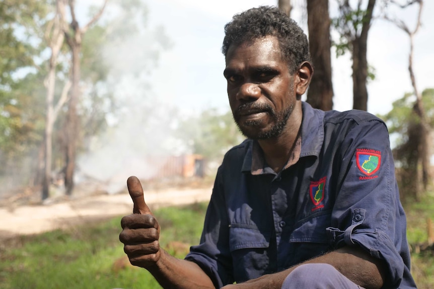 Indigenous ranger Dominic Bunduck is knelt in front of burning bushland and is giving a thumbs up.