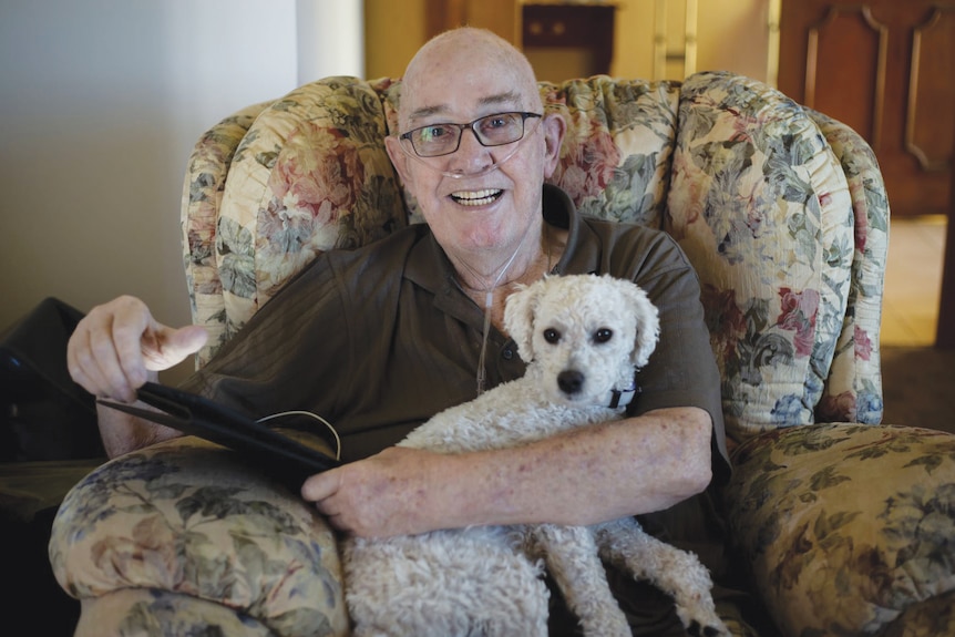 Older man sits with a small white dog on his lap in a floral armchair.