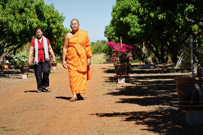 A woman and a man who is wearing orange robes walk past mango trees