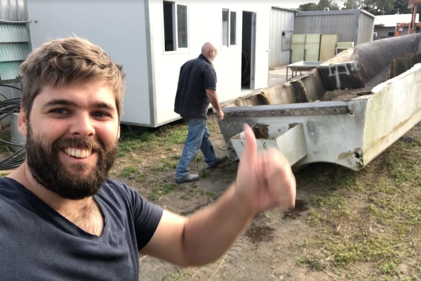 Adult son with dad in front of rundown boat in a backyard, smiling and thumbs up