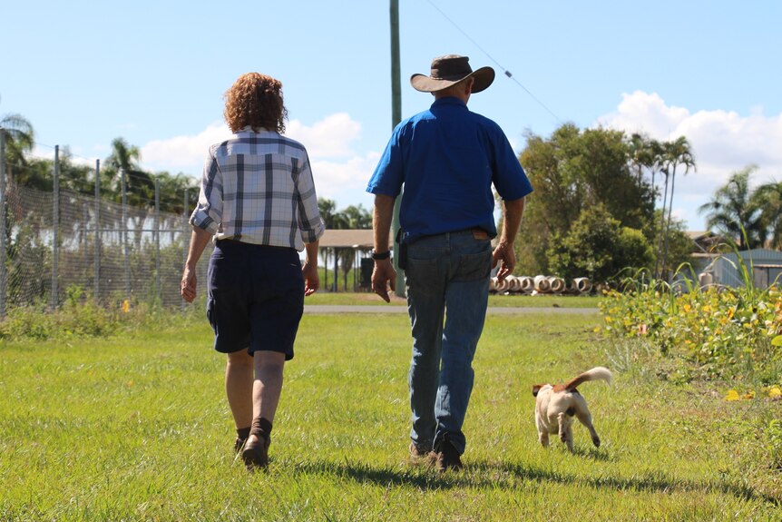 A man and a woman walk along grass with a little dog running at their heels