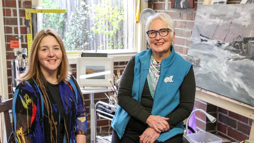 Two smiling women sit in a brick studio with paints, brushes and artworks in the background.