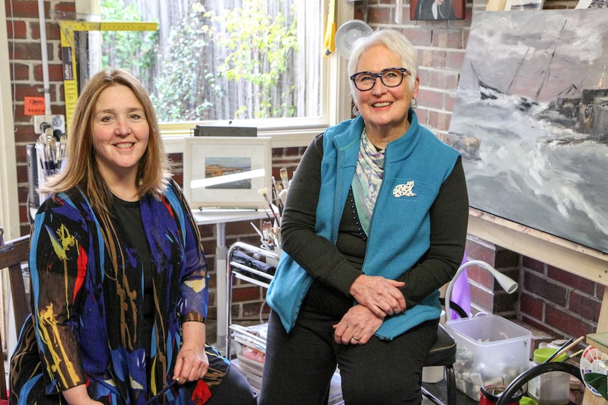 Two smiling women sit in a brick studio with paints, brushes and artworks in the background.