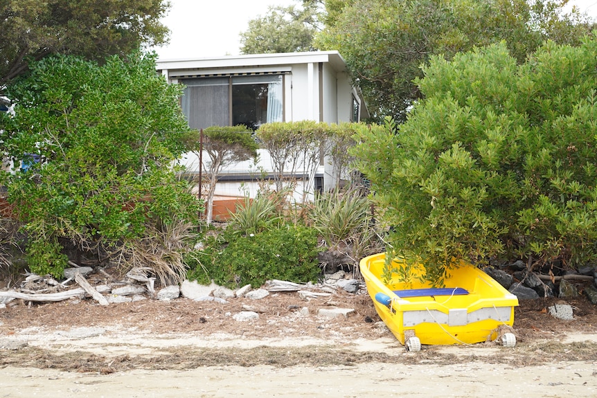 A small white house is seen from the beach with trees and a yellow boat in the foreground.