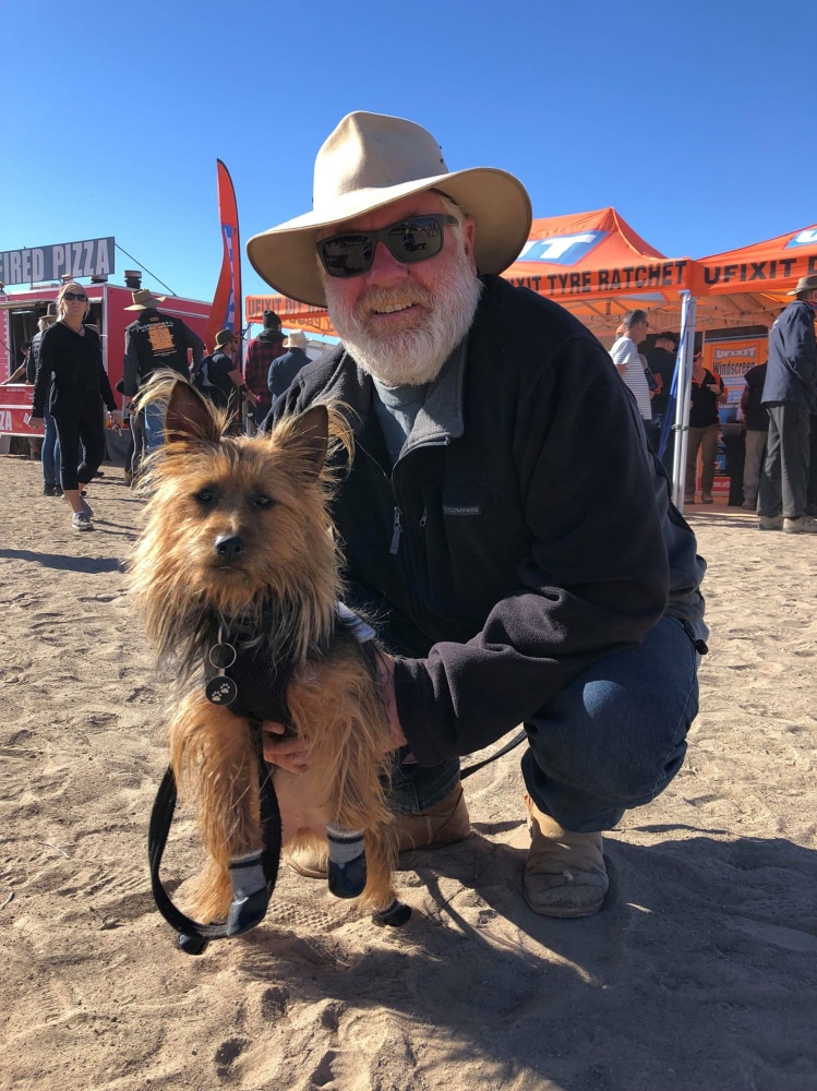 A man squats holding a small dog wearing boots, with festival stalls in the background.