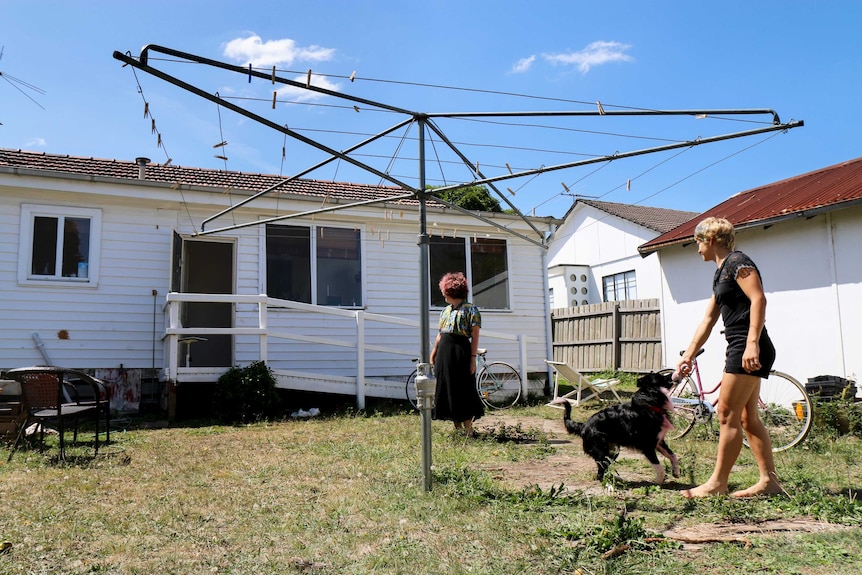 A blue sky, white weatherboard house with a hills-hoist in the foreground, and Sebastian and Jasmine standing barefoot on grass