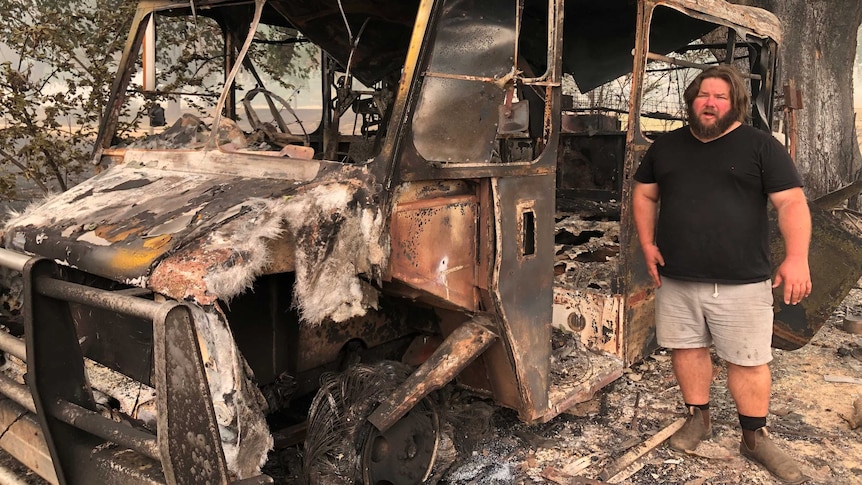 A man standing in front of the burnt ruins of a van or bus.