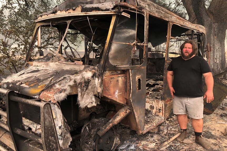 A man standing in front of the burnt ruins of a van or bus.