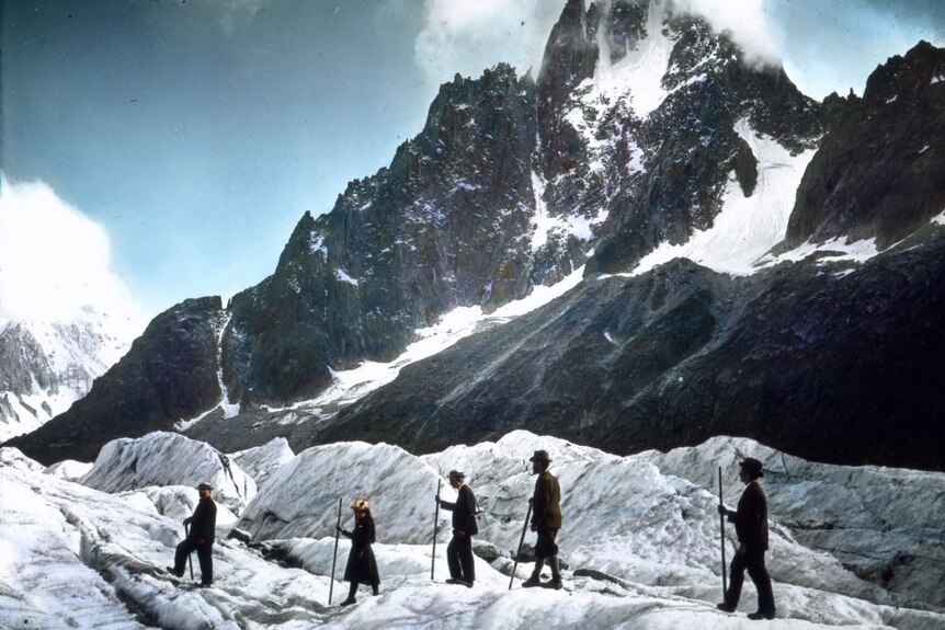 Four men and a woman, all dressed in old-fashioned clothes, hike across a glacier with a snow-topped mountain in the distance.