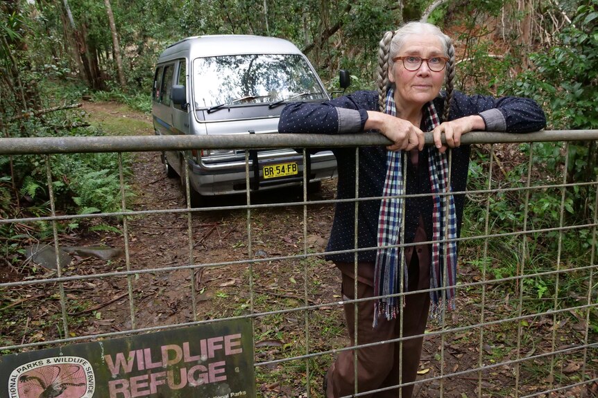 An older woman stands with her arms over a fence, surrounded by lush bushland