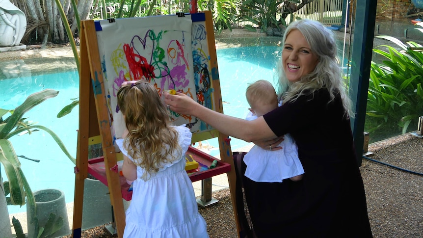 A young mother with holding an infant and painting with her toddler daughter outside with a pool in the background. 