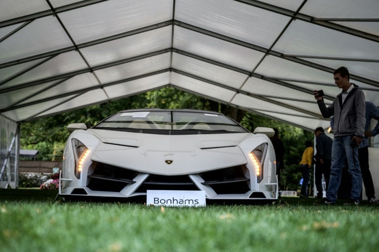 A white car is seen on grass and under a white shed. A man is seen to the right taking a picture of it.