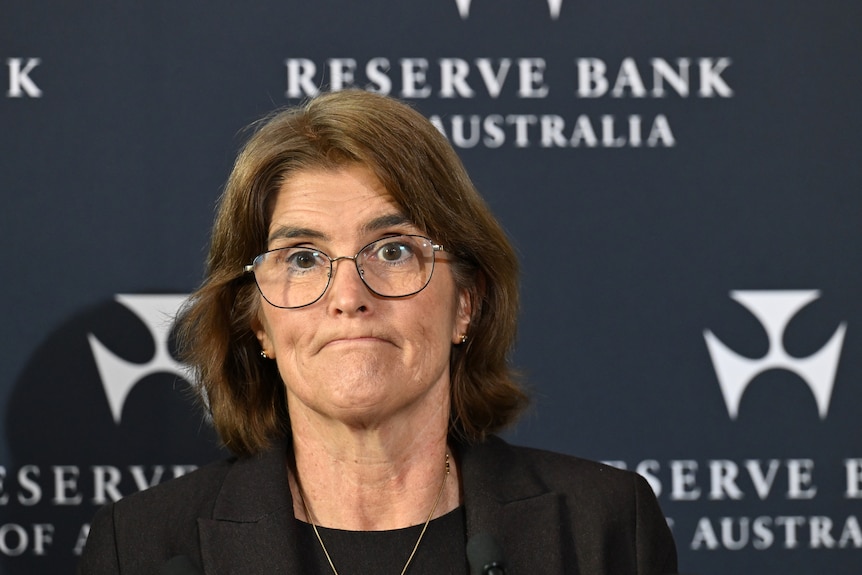 A woman with short brown hair and glasses speaks in front of a media wall that says "Reserve Bank of Australia".