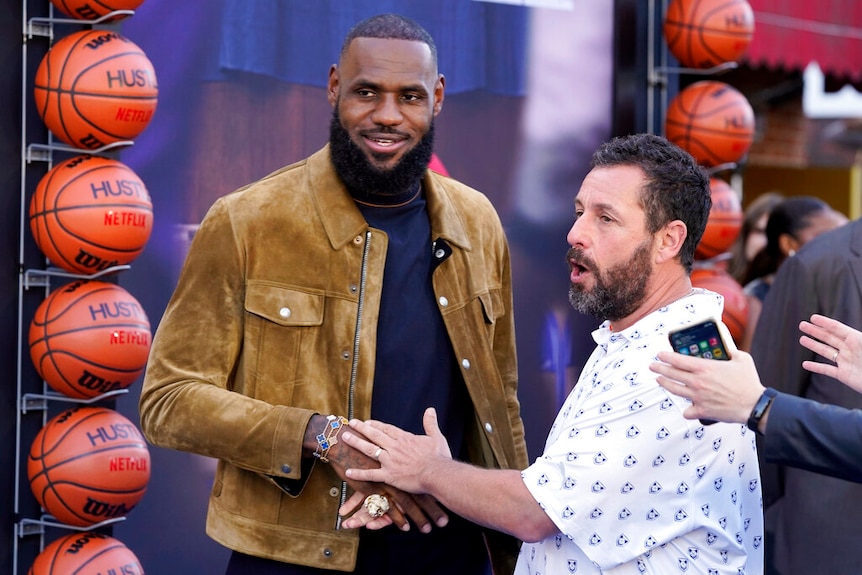 LeBron James shakes hands with Adam Sandler. Basket balls line the wall behind him. 