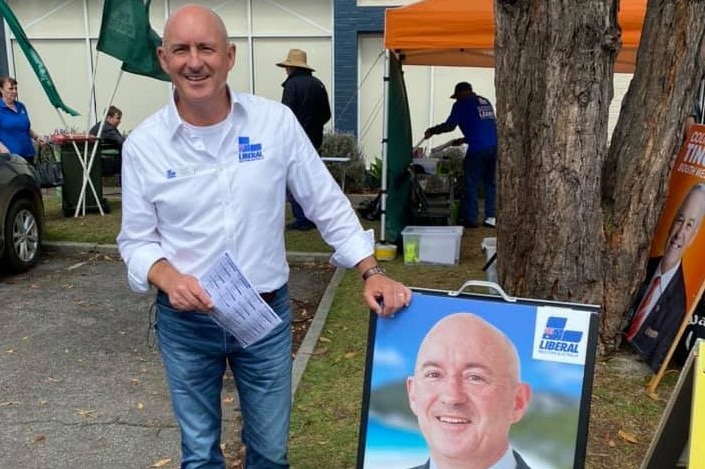 Scott wears a white shirt with a blue Liberal Party logo on the chest, standing next to a campaign sign.