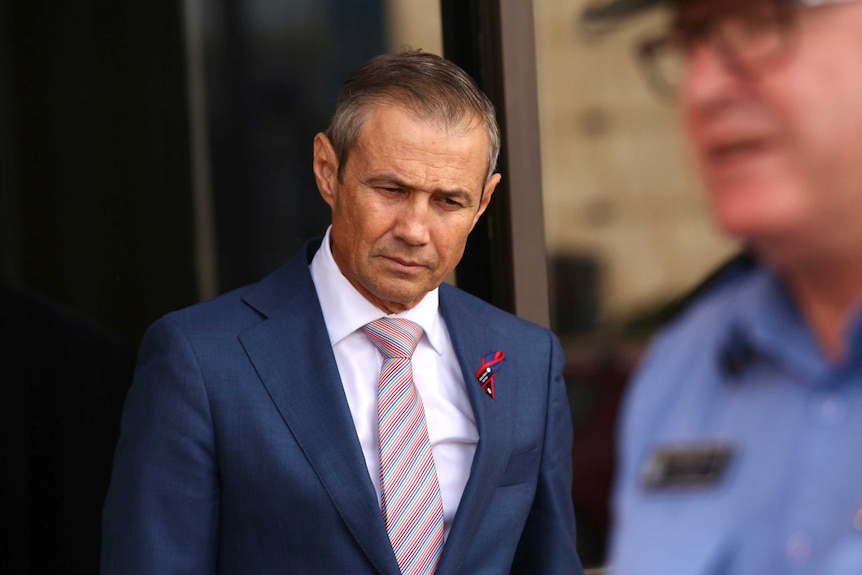 Roger Cook looks down with a serious face, while in the foreground out of focus Chris Dawson speaks.