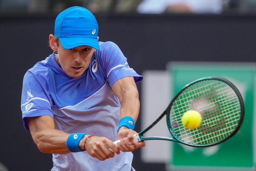 Professional male tennis player Alex de Minaur plays a two-handed backhand, hitting the ball, wearing a blue shirt and cap