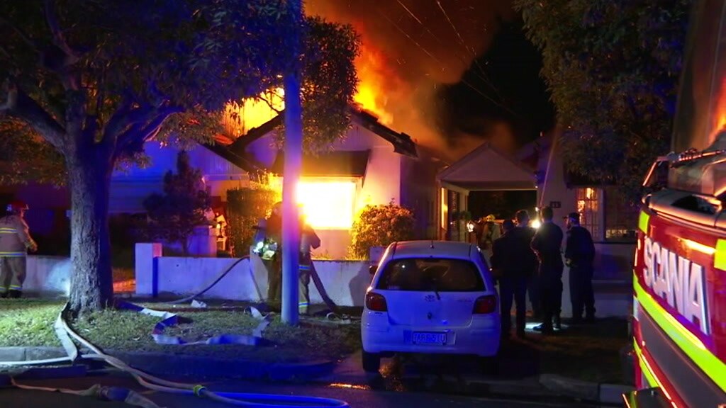 A house in flames with fire brigade hoses and car out the front.