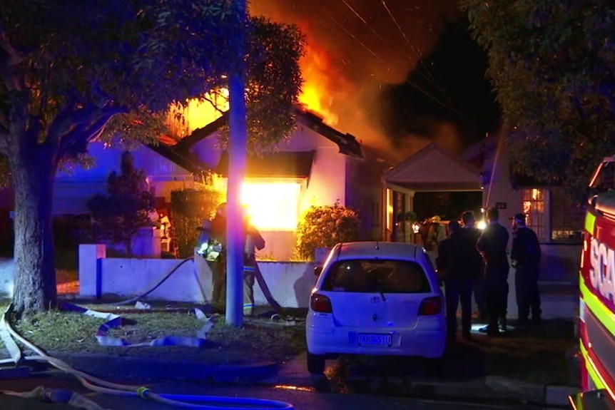 A house in flames with fire brigade hoses and car out the front.