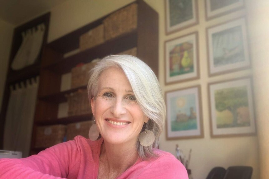 A woman with short white hair and big earrings smiles broadly with children's book illustrations on the wall behind her.