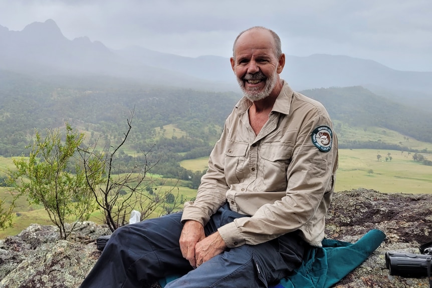 A man sitting on a rock, smiling at the camera with mountains in the background. 