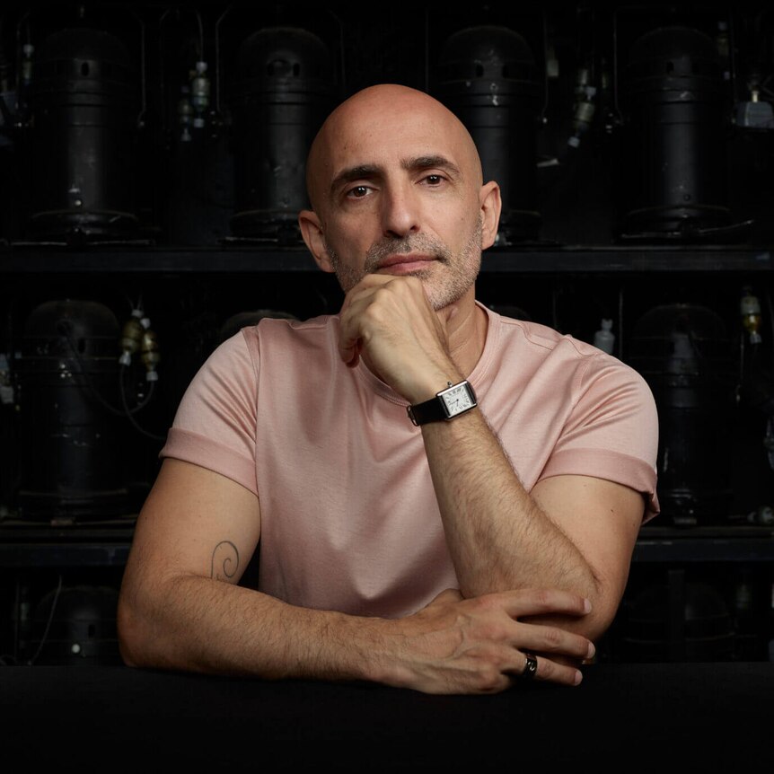 A portrait of Rafael Bonachela, in front of a shelf of stage lights, looking seriously at the camera with a hand on his chin.