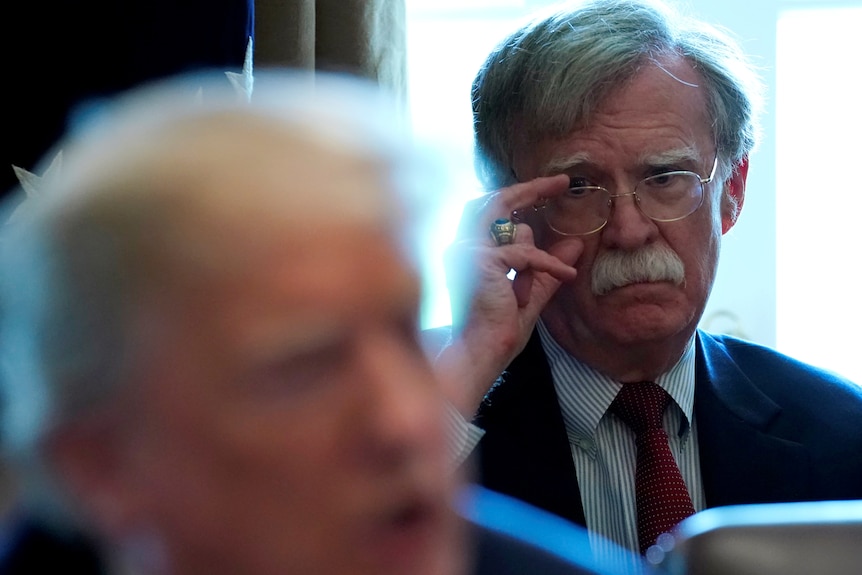 John Bolton sits behind Donald Trump. He is adjusting his glasses and frowning.