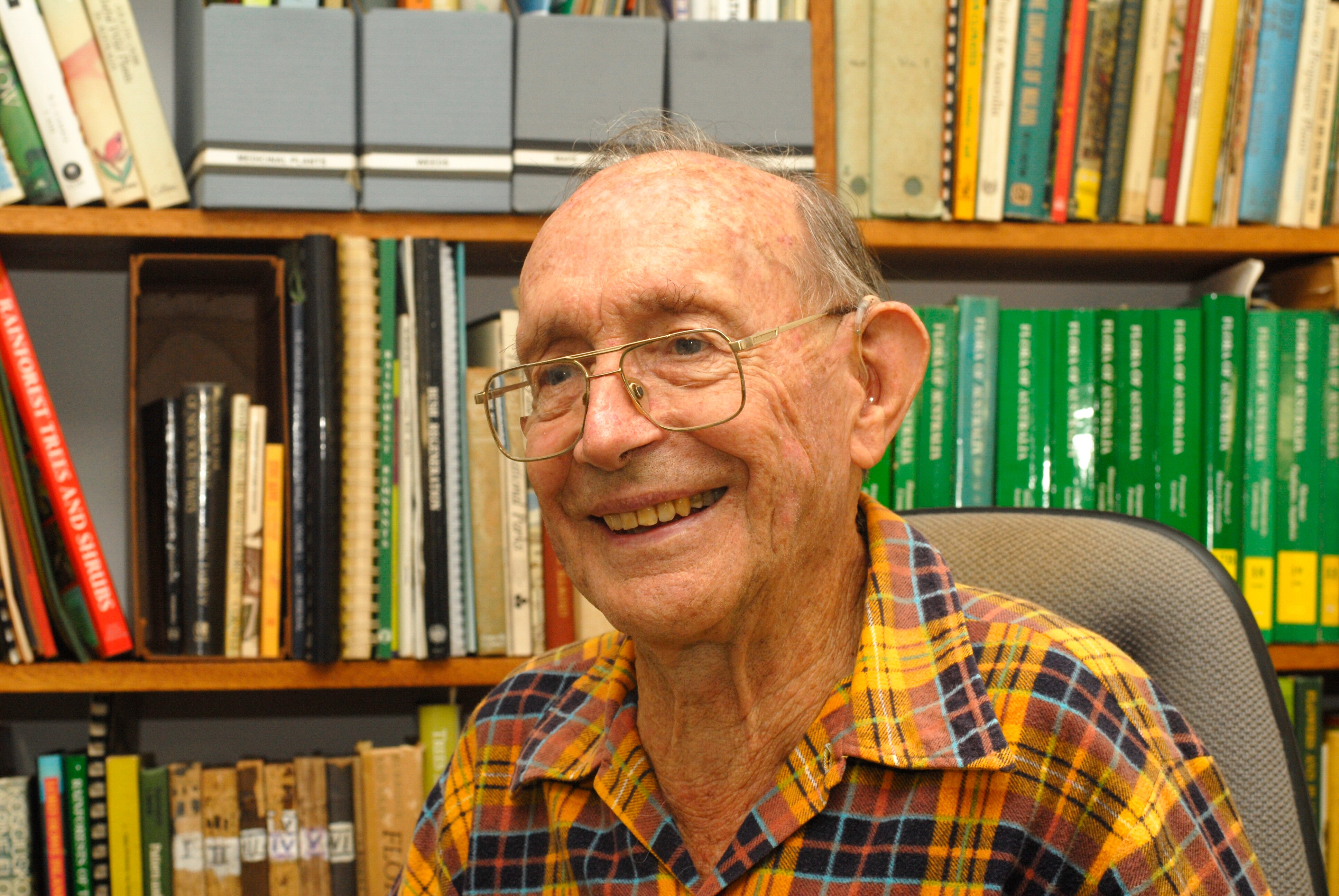 Elderly man wearing glasses and a yellow shirt sits in front of book shelf and smiles 
