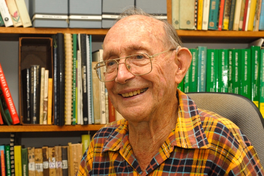 Elderly man wearing glasses and a yellow shirt sits in front of book shelf and smiles 