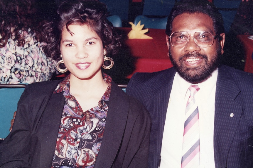 Narelda Jacobs, wearing a print shirt and hoop earrings, poses for a photo with her dad, who's wearing a suit and tie