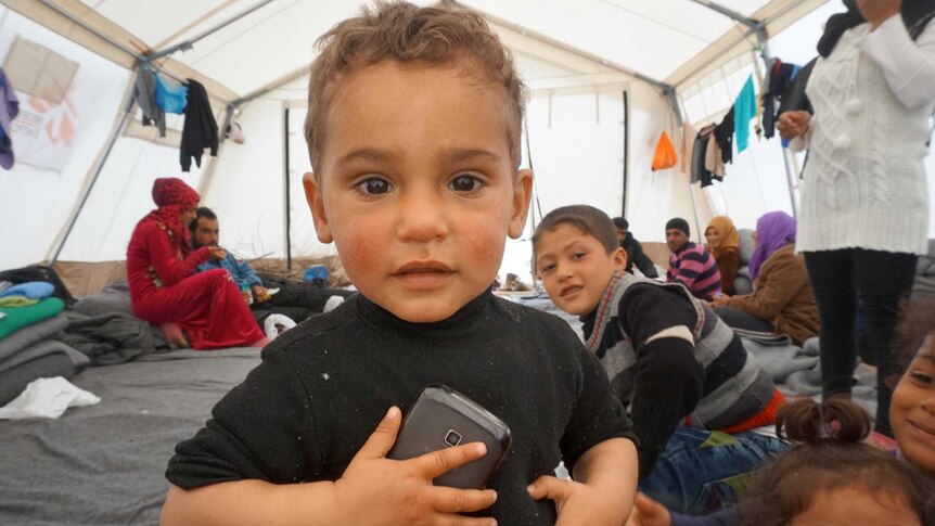 Upper body photo of a toddler holding a mobile phone to his chest as he stands in a tent.