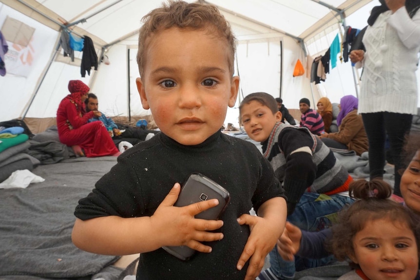 Upper body photo of a toddler holding a mobile phone to his chest as he stands in a tent.