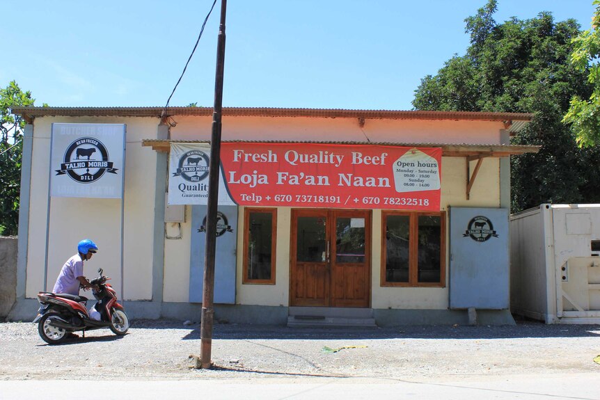 Man parks bike beside a new butcher shop in Dili, East Timor, with banners promoting Fresh Quality Beef in English and Tetum