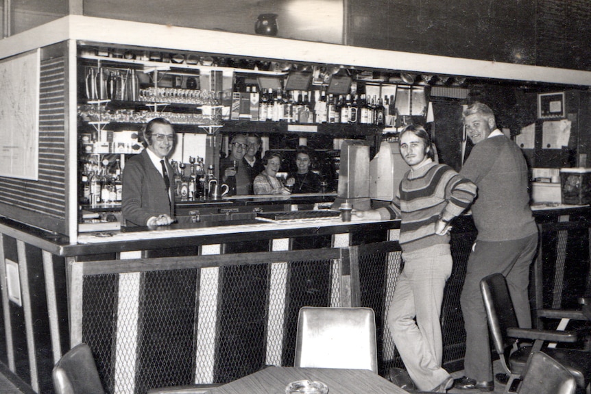 Two men leaning against the bar smile at the camera, as well as the barman. 