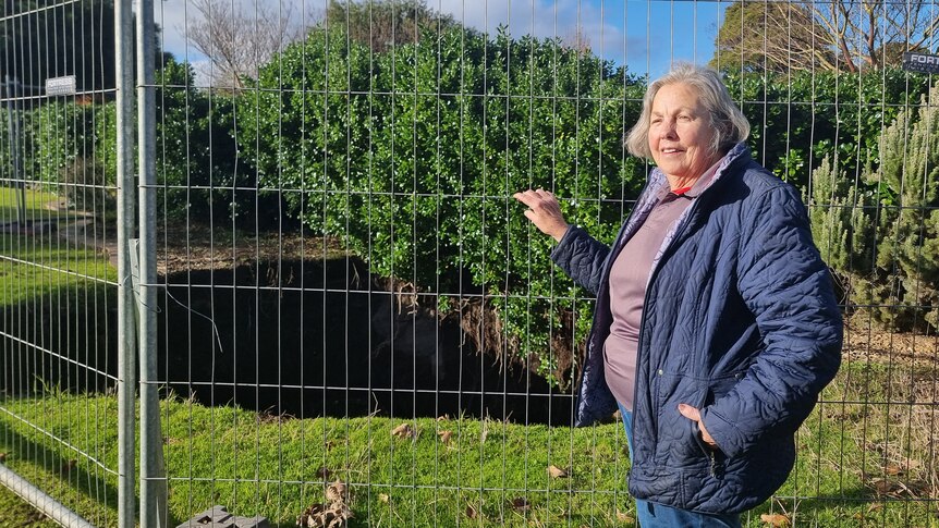 A lady holds on to a temporary fence with her left hand while looking at the camera, with a sinkhole behind the temporary fence.