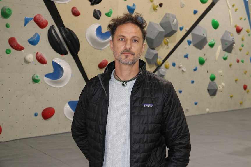Damien Hall wears a black jacket and stands in front of a rock climbing wall with coloured hand grips.