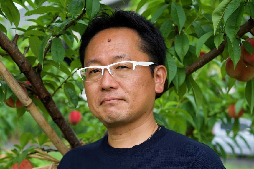 A man in white glasses and a blue t-shirt stands in front of a peach tree