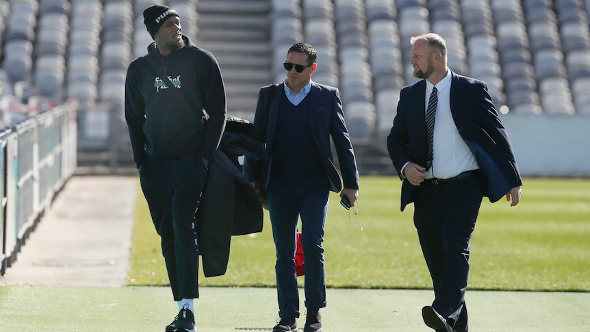 Usain Bolt walks inside a stadium wearing a tracksuit and hat next to two men wearing suits