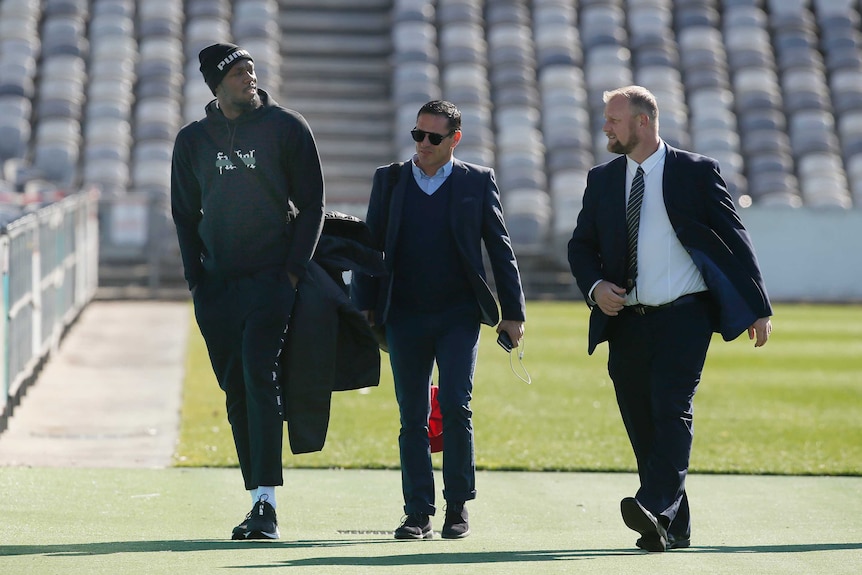 Usain Bolt walks inside a stadium wearing a tracksuit and hat next to two men wearing suits
