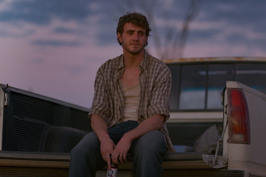 A white man in his 30s with brunette hair wearing a white singlet and checkered shirt sits in the back of a ute.