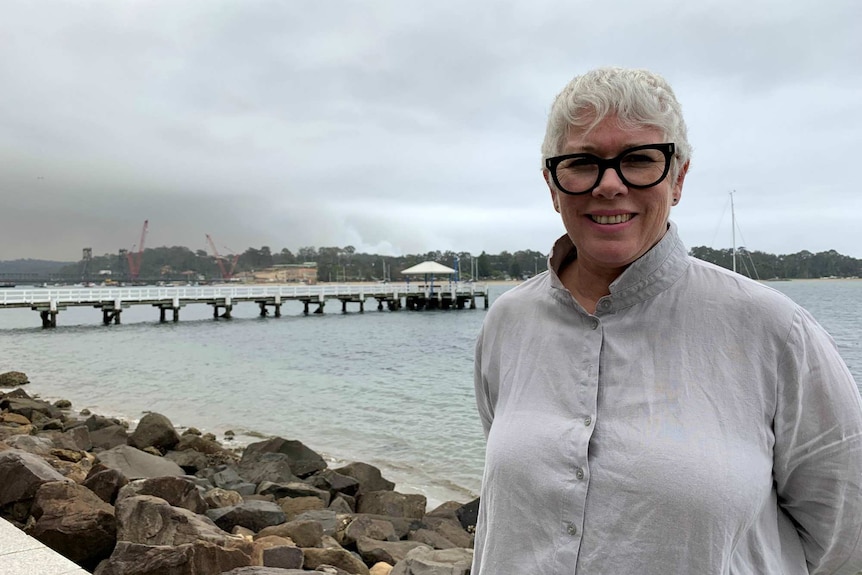 A woman wearing glasses stands with a pier and water in the background.