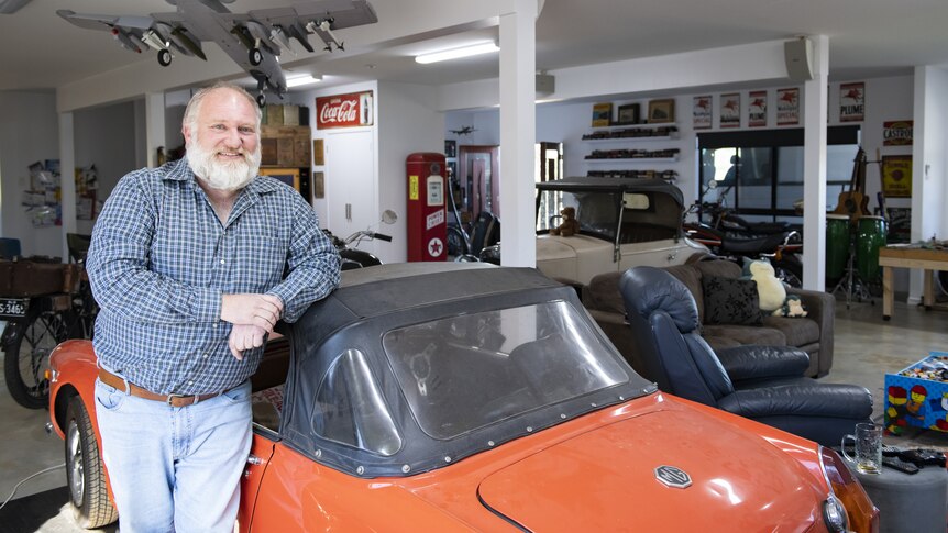 Mid shot of an older man with a short white beard leaning on the roof of an orange vintage car