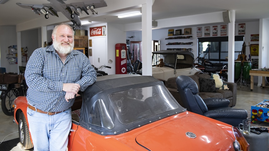 Mid shot of an older man with a short white beard leaning on the roof of an orange vintage car