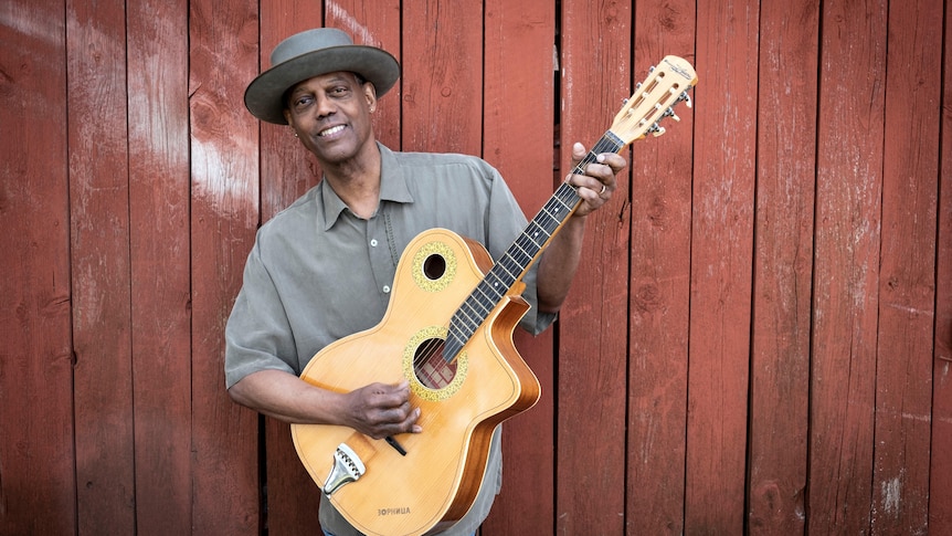 A smiling man in a wide brimmed hat, strumming an acoustic guitar