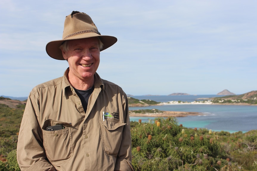A man in a wide brim hat standing in the bush with the ocean behind him. 