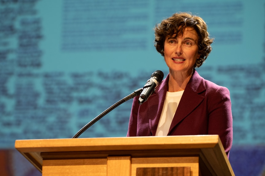 A mid-shot of Kate Chaney speaking into a microphone at a podium indoors, wearing a purple jacket and white top.