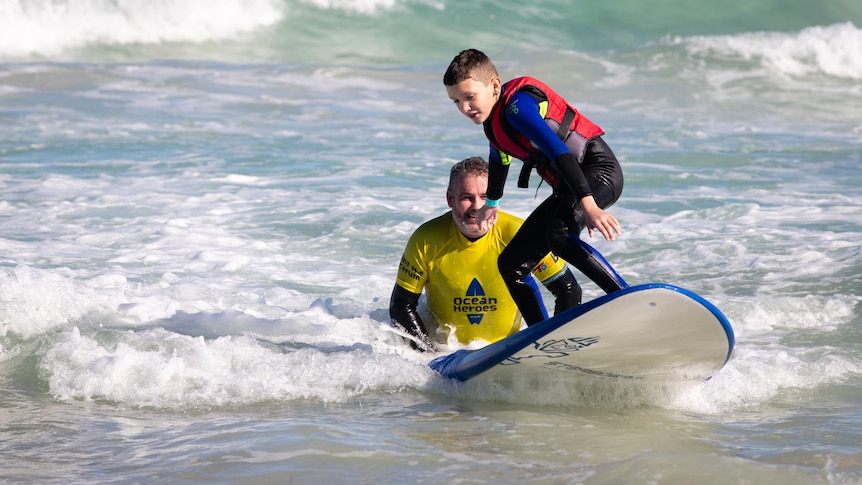 A young boy with a red life vest stands on a surfboard in whitewash, pushed by a man.
