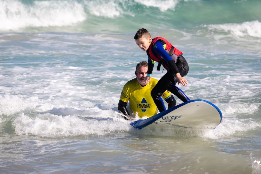 A young boy with a red life vest stands on a surfboard in whitewash, pushed by a man.