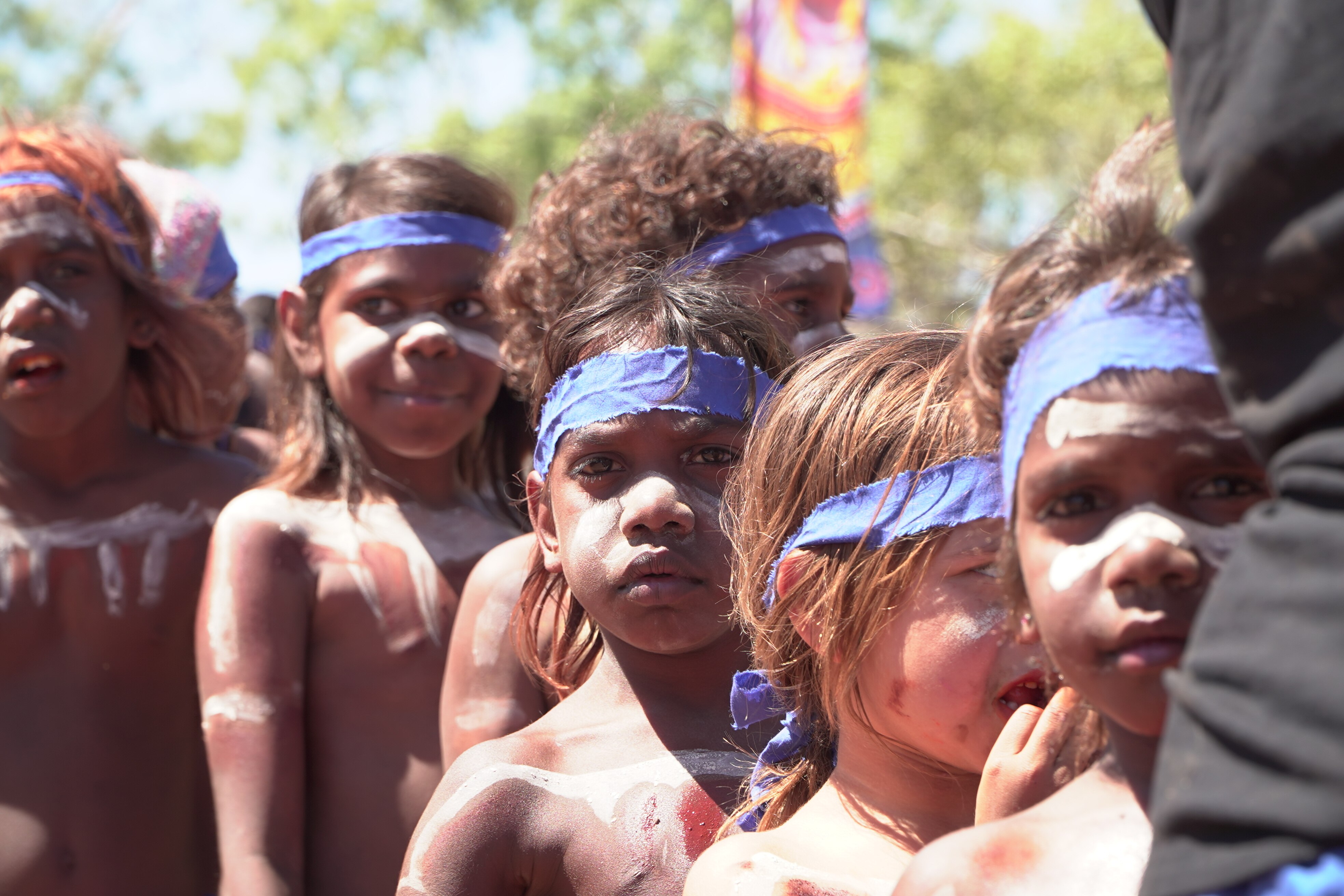 A group of young Aboriginal children with paint on their body, wearing blue and purple bandanas across their heads.