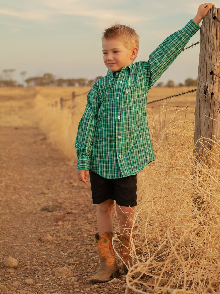 A young boy dressed in his best checked shirt and boots leans against a fence post.